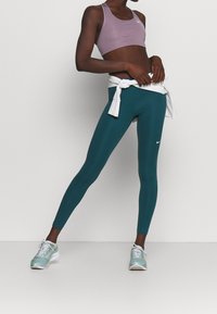 Persoon in paarse sportbh, teal leggings, witte vastgebonden sweatshirt en lichtblauwe sneakers die tegen een effen achtergrond staat.