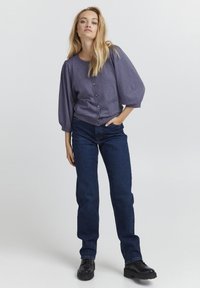 Lilla kootud kardigan puffvarrukatega, eesnööbiga sulgemine ja lapitaskud, kombineeritud tumedate siniste sirgete pükstega ja mustade kingadega.