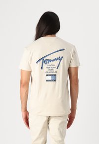 Lys beige t-shirt, korte ærmer, med et logo i denimtekstur i blå og bynavne trykt i sort på ryggen. Glat stofstruktur.