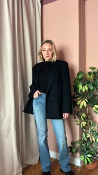 Νεαρή γυναίκα με ξανθά μαλλιά σε καρέ, φοράει μαύρο oversize σακάκι, μαύρη μπλούζα, ανοιχτό μπλε τζιν, στέκεται σε εσωτερικό χώρο δίπλα σε μπεζ κουρτίνα και φυτευτό φυτό.