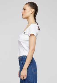 Bílý pruhovaný crop top s krátkými rukávy, s výstřihem do V a malým vyšitým logem na levé straně. Ležérní, přiléhavý design.
