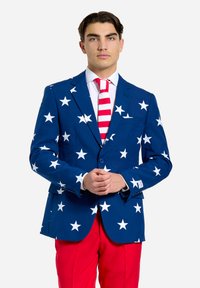 Blå blazer med store hvide stjerner, parret med røde bukser. Blazeren har et skræddersyet design og en enkelt knaplukning. Rød og hvidstribet slipse.