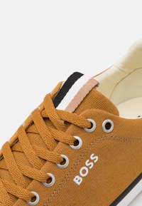 Primer plano de una zapatilla de lona marrón con cordones naranjas, ojales plateados, detalle de tela a rayas y el logotipo "BOSS" en blanco en el lateral.