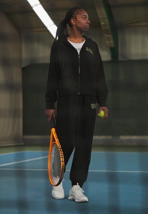 Felpa nera sportiva e pantaloni abbinati con loghi, che tiene in mano una racchetta da tennis arancione e una pallina da tennis verde, su un campo blu al chiuso.