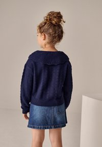 Marineblau gestrickter Pullover mit gewelltem Kragen und strukturierten Details, kombiniert mit einem Jeansrock mit einer Gesäßtasche. Rückansicht.