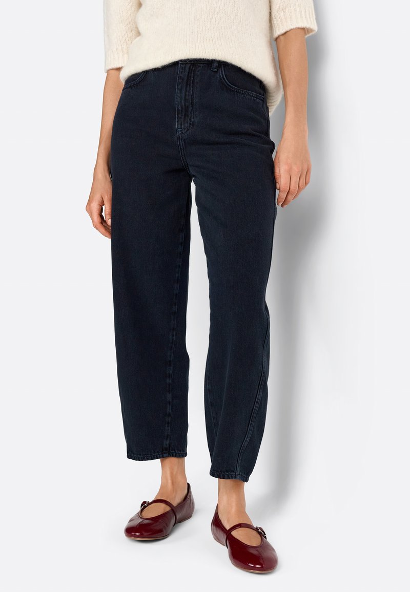 Jeans de mezclilla azul oscuro con cintura alta, corte holgado y piernas rectas, combinados con unos zapatos planos burdeos con un detalle decorativo.