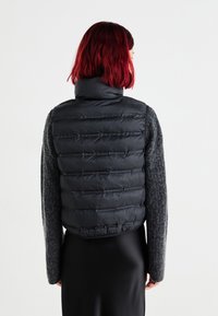 Zwarte puffer vest met gequiltde textuur, hoge kraag en een aansluitend ontwerp, gecombineerd met een grijs, wolachtig lange mouwen top en een zwarte rok.
