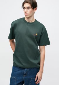 Tmavě zelené bavlněné tričko s volným střihem, kulatým výstřihem a malým vyšívaným logem na hrudi. Krátké rukávy a hladká textura.