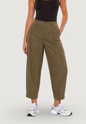 Persona che indossa pantaloni larghi corti verde oliva, top nero senza maniche e scarpe da ginnastica bianche, in piedi con una mano in tasca.