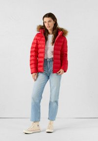 Giacca rossa imbottita con cappuccio foderato in pelliccia, caratterizzata da tessuto trapuntato. Abbinata a jeans di denim chiaro e sneakers beige alte.