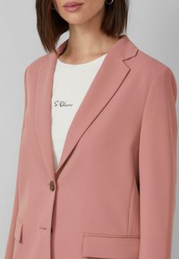 Vrouw in een stofroze blazer over een witte shirt met de tekst "S.Oliver", met een creolenoorbel en steil bruin haar, lichte grijze achtergrond.