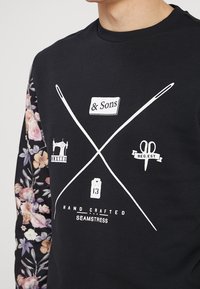 Sweat-shirt noir avec des manches à motif floral. Le devant présente des graphiques blancs d'outils de couture et des éléments de texte liés à l'artisanat.