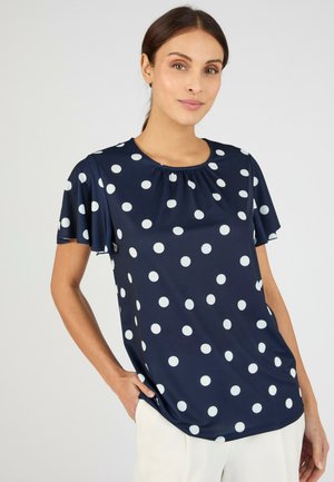 Mujer que lleva una blusa azul marino con lunares blancos y mangas cortas acampanadas, combinada con pantalones de color claro, parada con una mano en el bolsillo.