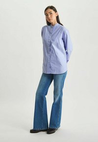 Chemise boutonnée à manches longues bleu clair avec un col mandarin, associée à un jean en denim bleu évasé et des chaussures noires.