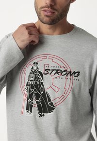 Camisa de manga larga gris con un gráfico estilizado de un personaje sosteniendo un sable de luz rojo, rodeado de un emblema circular y texto.