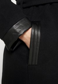 Manteau noir en tissu texturé avec un poignet en cuir et une poche latérale, mettant en avant une finition lisse et des détails matelassés.