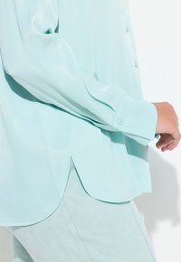 Lyseblå turkis bluse lavet af glat stof. Den har knapper foran, lange ærmer og en buet kant med en slidsdetalje.