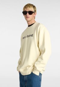 Sudadera de color crema con el texto negro "dile a un amigo." en el frente, llevada sobre una camiseta oscura, combinada con jeans azules y gafas de sol oscuras.