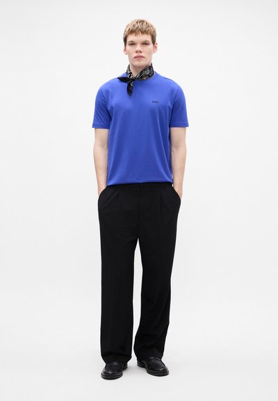T-shirt en coton bleu à manches courtes pour homme avec un logo sombre, associé à un pantalon noir plissé et une écharpe noire à motifs autour du cou.