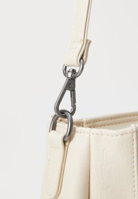 Détail d'un sac en cuir crème avec une boucle en métal, une bandoulière robuste et des coutures visibles. Surface texturée sans motifs ni impressions.