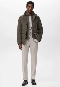 Olivengroene pufferjack met een hoge kraag, knoopsluiting, gedragen over een lichtgrijs pak. Tappered beige broek en zwarte schoenen maken de look compleet.