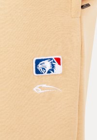 Tessuto beige con una piccola toppa ricamata raffigurante una testa di tigre bianca che ruggisce su uno sfondo blu e rosso, con un logo astratto bianco sottostante.