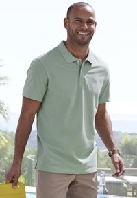 Polo shirt verde chiaro con colletto e maniche corte, realizzato in tessuto morbido. Pantaloni corti beige, con un oggetto giallo in mano.