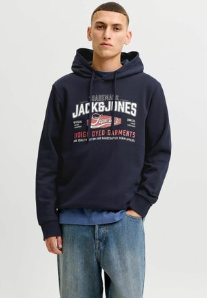 Junger Mann trägt einen marineblauen Jack & Jones Hoodie und hellblaue Jeans, steht mit einer Hand in der Tasche vor einem einfarbigen Hintergrund.