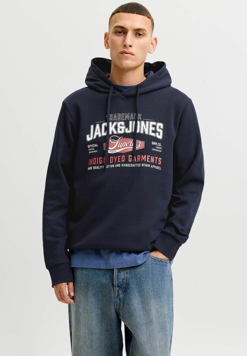 Jeune homme portant un sweat à capuche bleu marine Jack & Jones et un jean bleu clair, debout avec une main dans la poche devant un fond uni.