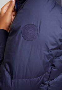 Mörkblå pufferjacka med en slät yta, som har en cirkulär gummilogo på vänster axel med texten "JUST THE PRODUCT."