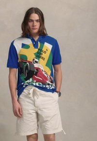 Kortermet polo skjorte i blå med fargerik racinggrafikk og logo. Kombinert med hvite shorts. Stoffet ser glatt og lett ut.