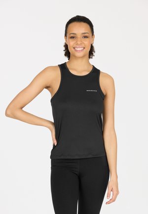 Femme souriante aux cheveux bouclés, vêtue d'un débardeur de sport noir et de leggings, debout avec une main sur la hanche sur fond blanc.