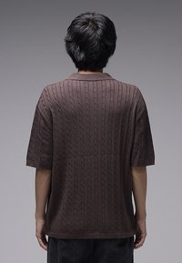 Καφέ κοντομάνικο πουλόβερ με υφή καλωδιωμένης πλέξης και γιακά πουκάμισου. Η πίσω πλευρά έχει ομαλή, μονή περιοχή με ελάχιστες λεπτομέρειες.