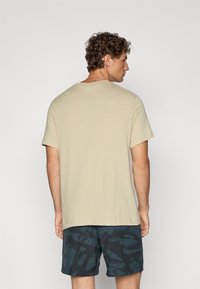 Beige bomulds t-shirt med rund hals og korte ærmer, parret med mørke mønstrede shorts. Synlige sømme og enkelt design.