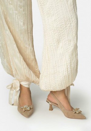 Tacones slingback puntiagudos beige con grandes adornos de cristal, combinados con pantalones crema texturizados y recogidos, atados en los tobillos.