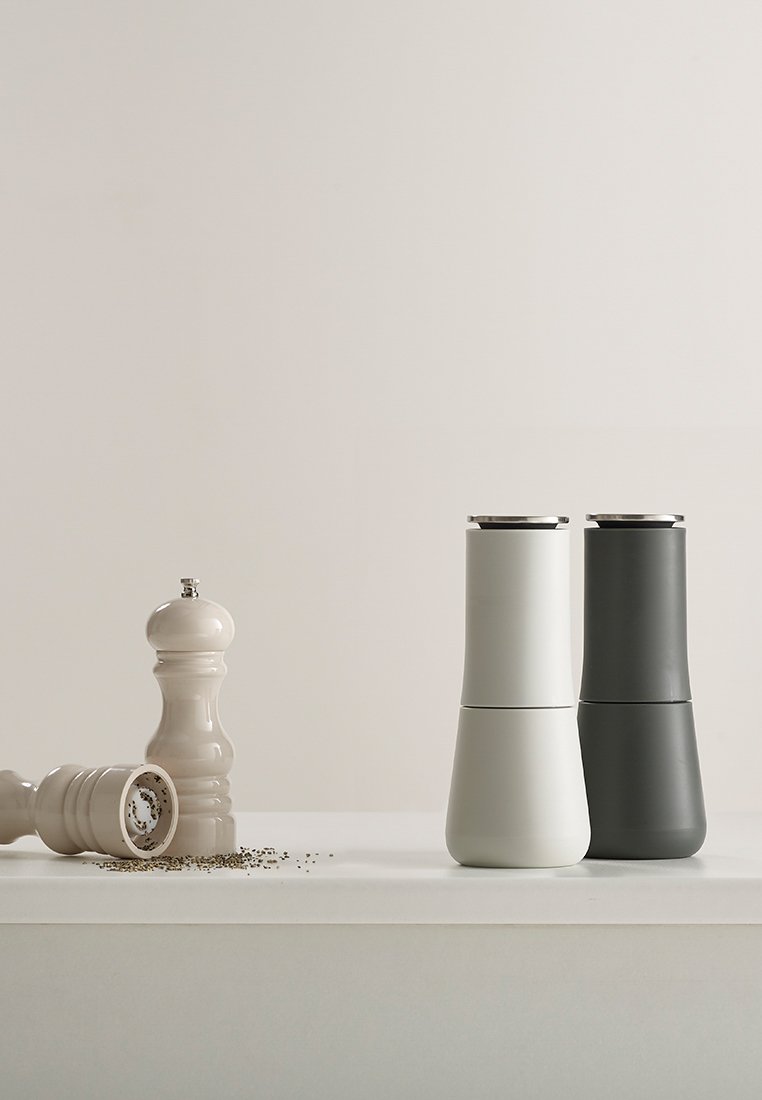 Witte en donkergrijze keramische zout- en pepermolens, cilindrisch met een gladde textuur, voorzien van een metalen bovenkant en peper die over het oppervlak is gestrooid.