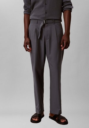 Hombre que lleva una camisa gris oscuro abotonada y pantalones plisados a juego con cinturón negro con hebilla y sandalias negras sobre fondo gris.