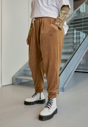 Osoba nosi brązowe, teksturowane spodnie dresowe Nike, białe sznurowane buty z czarnymi podeszwami oraz białą koszulę z długimi rękawami w zielono-brązowy wzór.