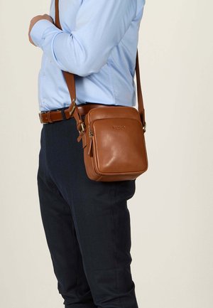 Brown leather crossbody bag with a square shape, adjustable strap, front zipper pocket, and embossed logo detail.