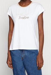 Hvid bomulds t-shirt med korte ærmer, der har en guldfarvet halsudskæring og trykt tekst "en lomme fuld af solskin" i en dekorativ skrifttype.