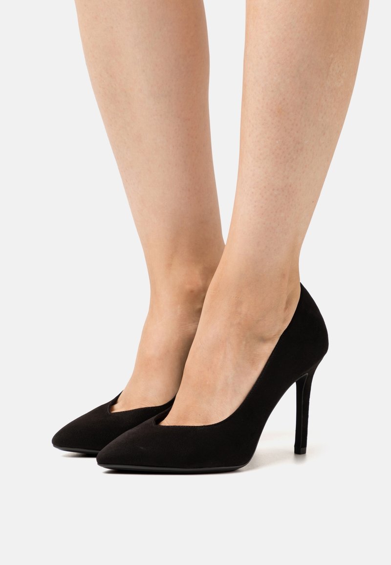 Jambes portant des escarpins noirs à bouts pointus avec talons stilettos sur un fond blanc.
