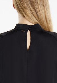 Schwarze Bluse mit hohem Kragen, Schlüssellochöffnung auf der Rückseite und zwei Knopfverschlüssen, aus glattem Material und minimalistischem Design.