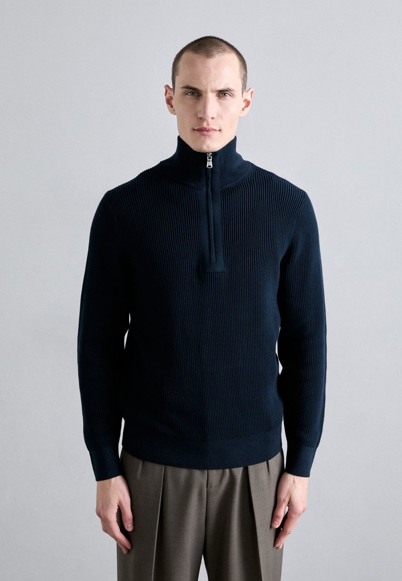 Marineblaue, gerippte Strickjacke mit hohem Kragen und einem halben Reißverschluss. Kombiniert mit grauen, gefalteten Hosen. Einfarbiger Hintergrund.