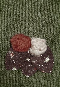 Il tessuto lavorato a maglia di un verde scuro presenta un abbellimento tessile con tre fiori di stoffa: due in lana marrone e beige, e uno con una stampa floreale scura.