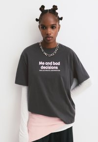 Jonge vrouw met gedraaide bantu knopen, gekleed in een donkergrijze t-shirt met de tekst "Ik en slechte beslissingen, de Ultieme Cross-over" en gelaagde kleding.