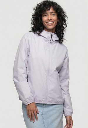 Lächelnde Frau mit lockigem Haar trägt eine hellviolette Jacke mit Reißverschluss und einen hellblauen Jeansrock vor einfarbigem Hintergrund.