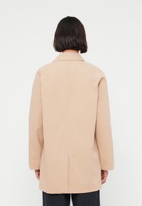 Μπεζ παλτό με ευθύ κόψιμο, μονή πίσω σχισμή και γιακά με χαραγή. Κατασκευασμένο από λείο ύφασμα, διαθέτει λιτό σχεδιασμό και μακριά μανίκια.