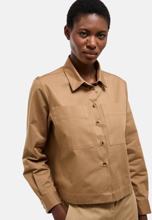 Femme aux cheveux courts portant une chemise beige boutonnée avec deux poches poitrine, debout devant un fond clair uni.