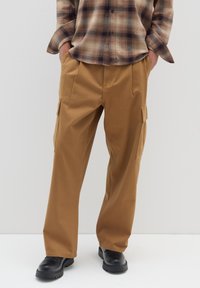 Braune Cargo-Hosen mit Seitentaschen, schlank geschnitten, aus glattem Stoff, kombiniert mit einem karierten Hemd und schwarzem Schuhwerk.