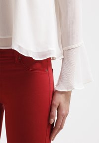 Blusa bianca plissettata con maniche a campana, abbinata a jeans rossi aderenti. La blusa presenta un delicato pizzo decorativo ai polsini.
